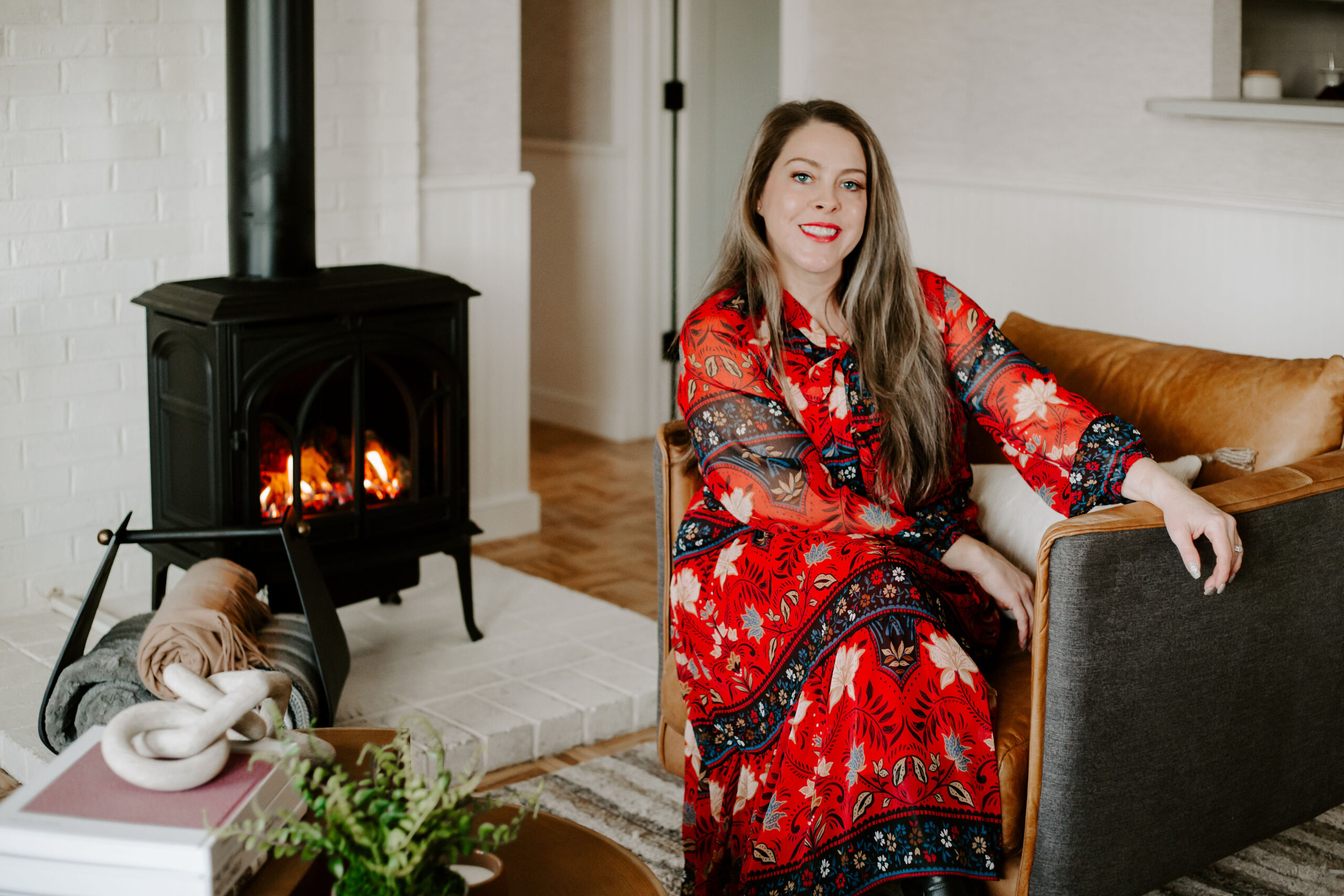 Interior designer, Emilie Kyle sits in leather chair in front of a wood burning stove next to an accessorized coffee table