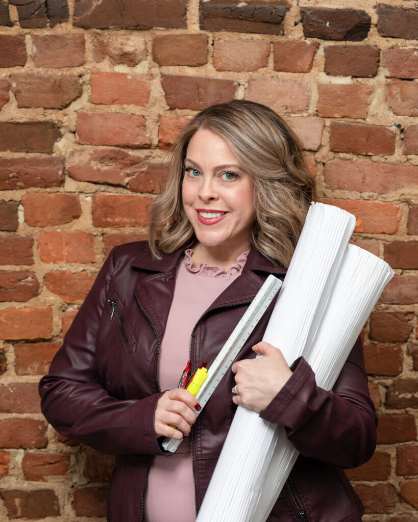 Alexandria Interior Designer, Emilie Kyle standing in front of a brick wall holding rolled architectural plans, a scale, a red pen, and a highlighter
