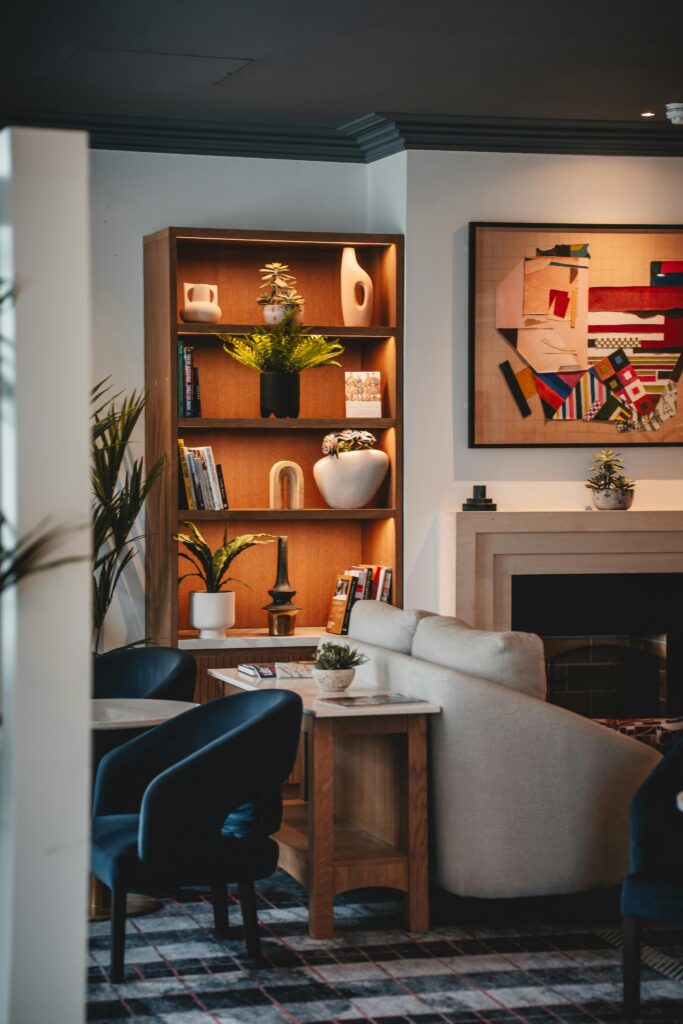 view of living room from another room, walls are white, ceiling and crown trim are a dark inky blue-charcoal color, shows styled bookcase with a palm plant on the left, fireplace on the right, wood base with marble top console table with plant and magazines on top, small linen upholstered curve back loveseat facing right with higher back cushions, two dark teal chairs facing left in front of console, stepped stone fireplace surround with colorful abstract mixed media art above, rug is navy and light teal stripe with a thin pink stripe running perpendicular